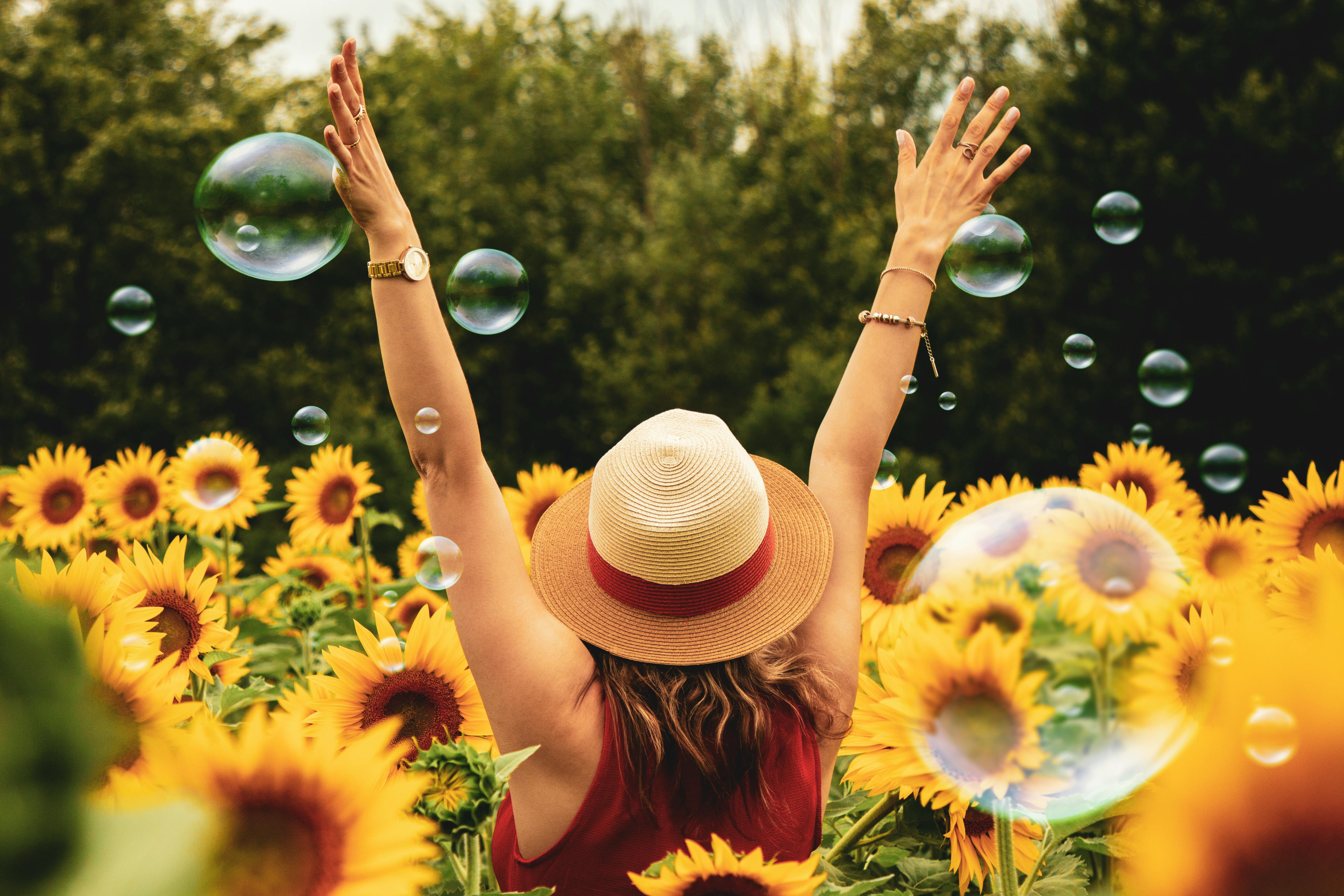 Back of woman in a field of sunflowers. Her arms are in the air and she's wearing a red tank top and straw hat. There are bubbles in the air.