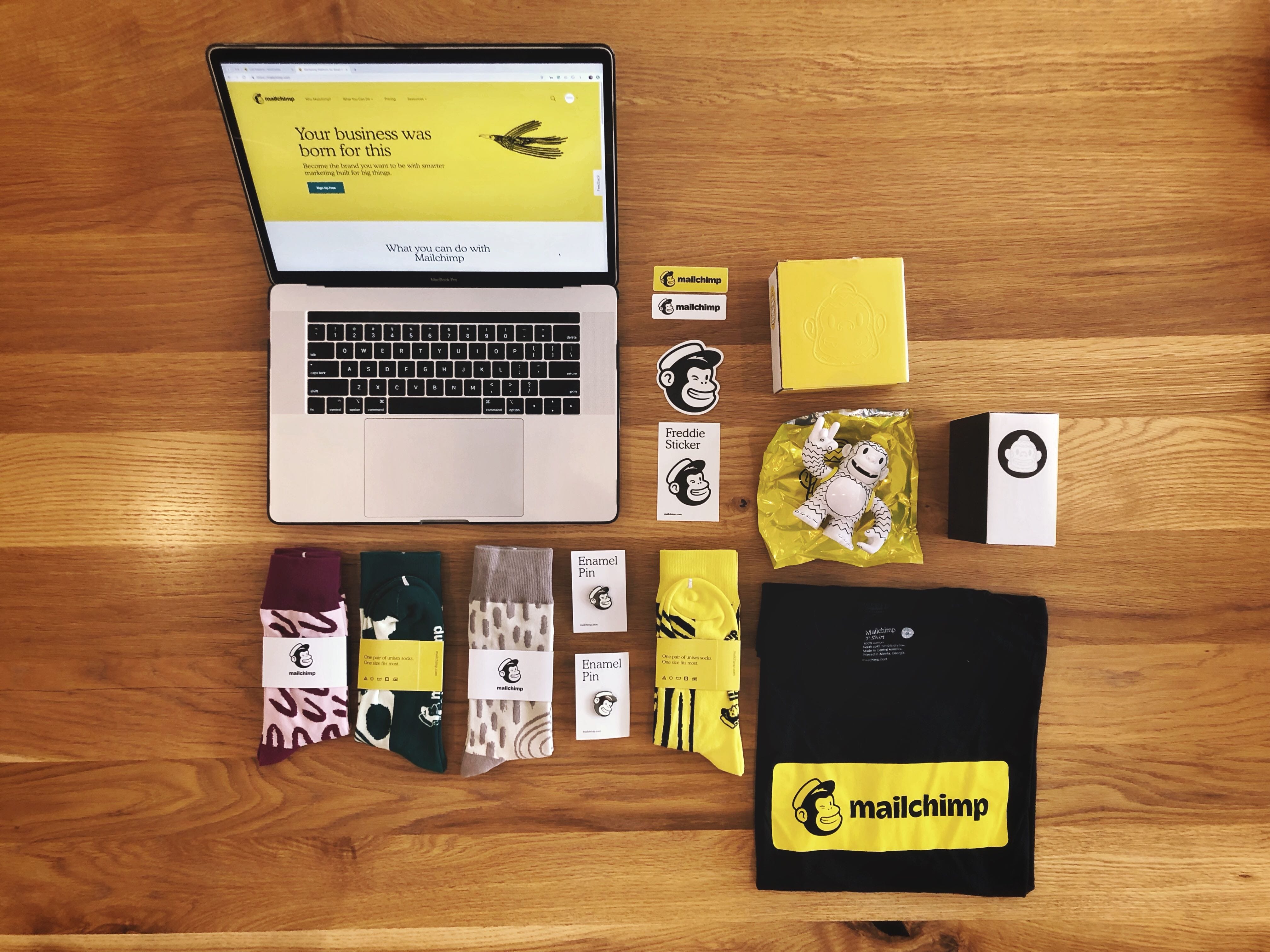 11 graphic design principles to elevate your brand 4 a computer, pins, stickers and a t-shirt lay on a table—all branded with the signature yellow and black Mailchimp logo